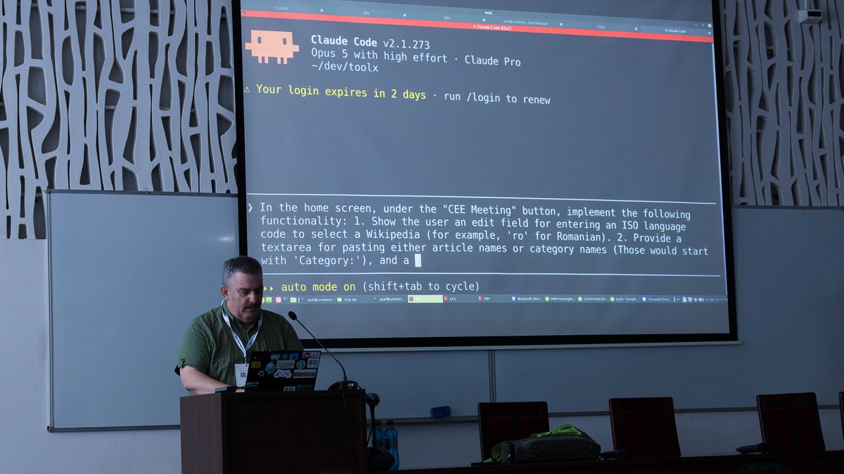 Bir konferans salonunda dev perdeye yansıtılmış Anthropic Claude Code terminal ekranı ve kürsüde dizüstü bilgisayarıyla konuşan katılımcı