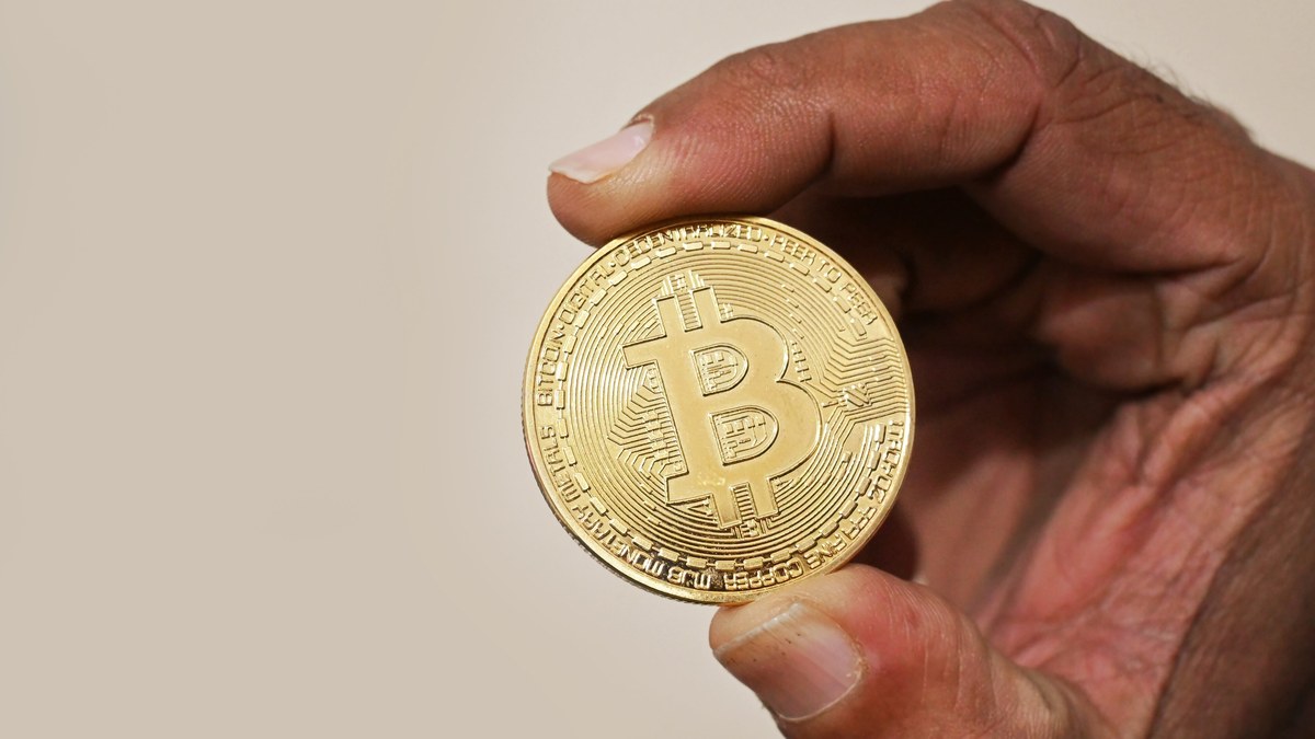 Açık renk zemin önünde iki parmak arasında tutulan altın renkli Bitcoin hatıra parası (temsili görsel, 2023)