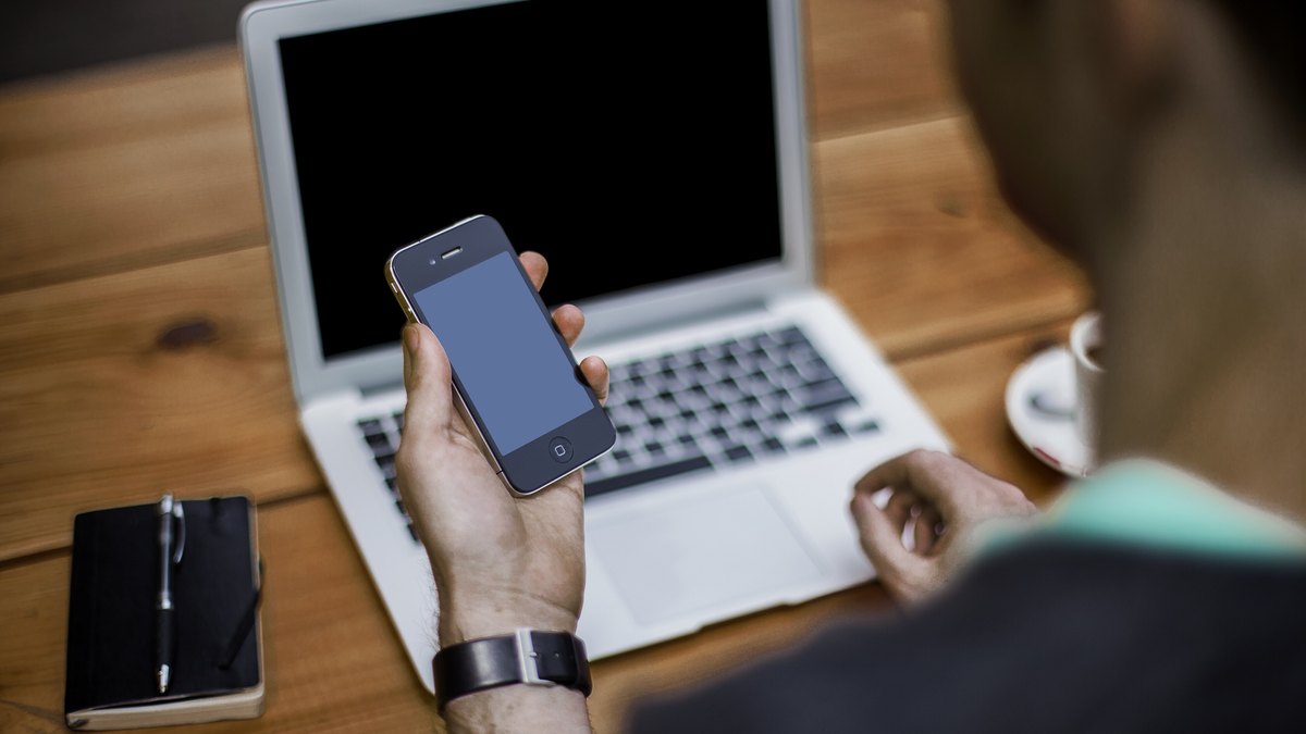 Dizüstü bilgisayar başında elindeki akıllı telefona bakan kişi; web girişinde telefonla ikinci doğrulama adımını simgeleyen temsili fotoğraf (e-Devlet Kapısı ekranı değildir)