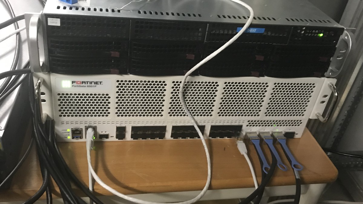 Ağ odasında kablolanmış, ön yüzünde Fortinet FortiGate 6501F yazan beyaz güvenlik duvarı cihazı ve üstünde rafa monte sunucu
