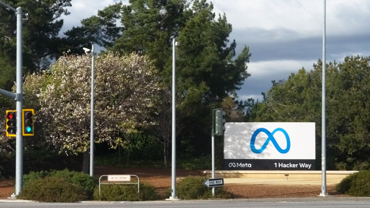 Meta'nın Menlo Park'taki genel merkez girişinde, ağaçlar ve trafik ışıkları arasında Meta logolu 1 Hacker Way tabelası