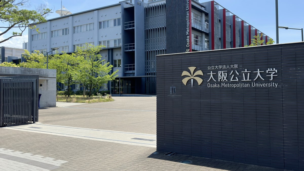 Osaka Metropolitan Üniversitesi Nakamozu kampüsünün girişinde 大阪公立大学 Osaka Metropolitan University yazılı tabela ve arkasındaki üniversite binası